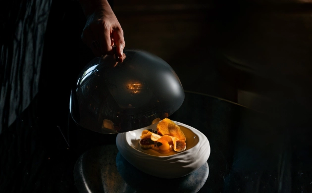 A plate with orange-colored food is served on a white dish, while a hand lifts a serving cloche to reveal the dish.