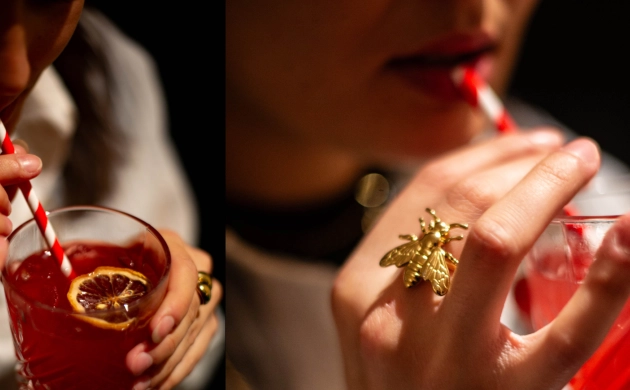 Mulher a beber um cocktail vermelho com palhinha às riscas, detalhe aproximado do copo e anel dourado em forma de abelha.