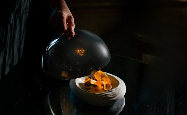 A plate with orange-colored food is served on a white dish, while a hand lifts a serving cloche to reveal the dish.