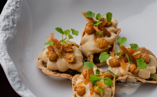A plate with orange-colored food is served on a white dish, while a hand lifts a serving cloche to reveal the dish.
