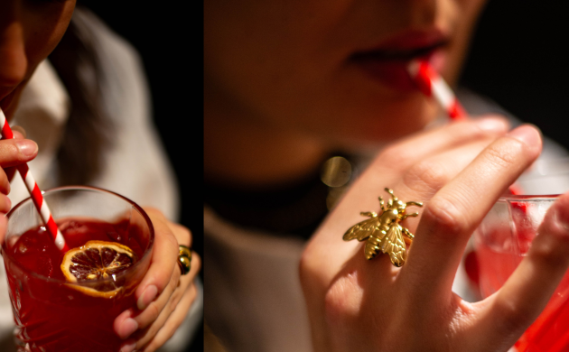 Mulher a beber um cocktail vermelho com palhinha às riscas, detalhe aproximado do copo e anel dourado em forma de abelha.